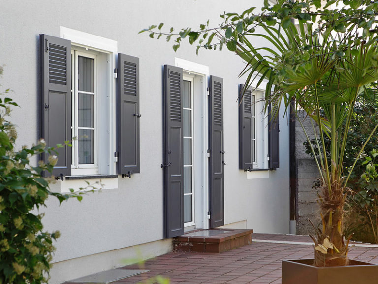 isol&plus-volets_battants_aluminium Volets battants en aluminium gris foncé sur une maison moderne en crépi gris clair