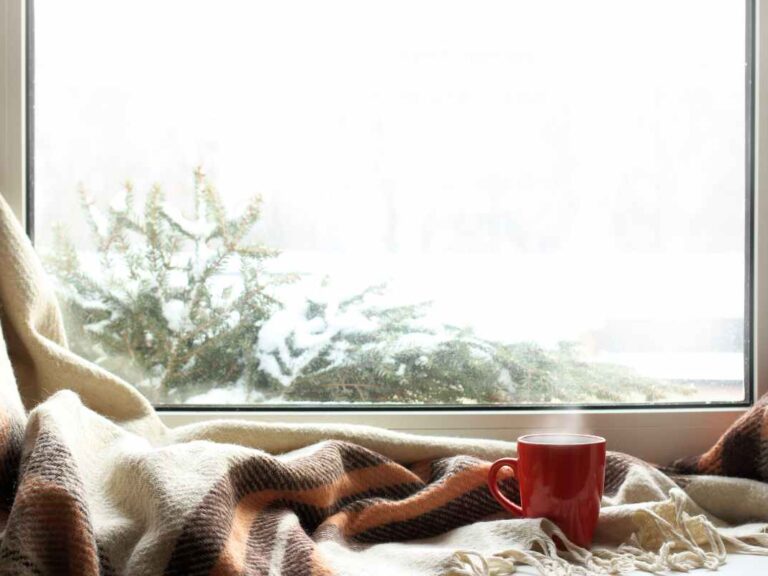 Fenêtre avec châssis en PVC blanc donnant sur un paysage enneigé et avec un plaid et une tasse rouge devant